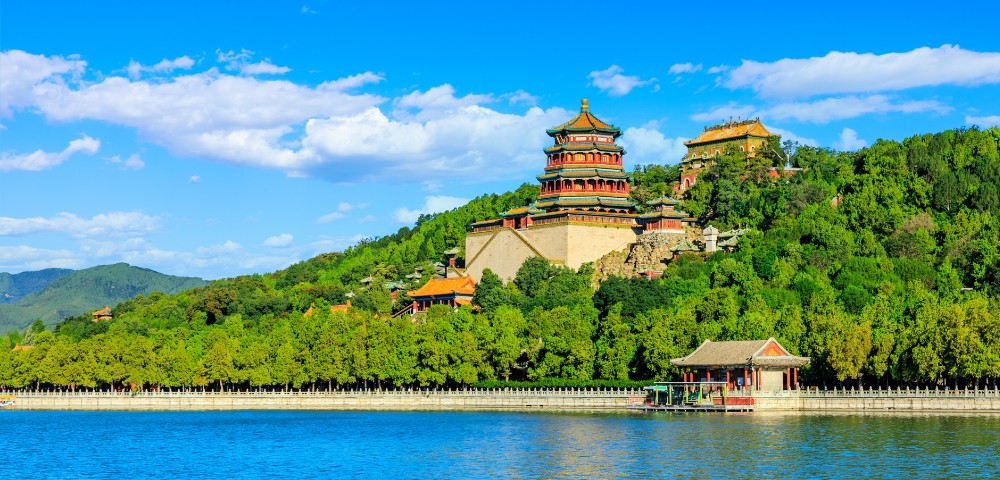 A traditional Chinese building with layered roofs sits on a lush green hillside above a calm blue lake under a clear, bright blue sky.