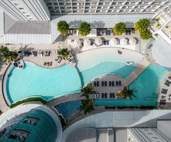 Aerial view of a luxurious resort pool area with curved, turquoise pools, lounge chairs, umbrellas, and palm trees. The scene feels relaxing and inviting.