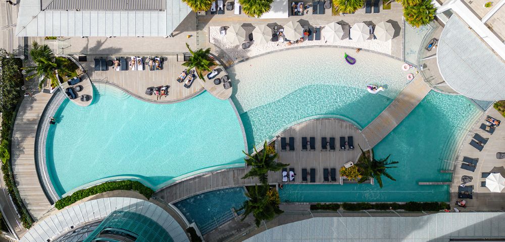 Aerial view of a luxurious resort pool area with curved, turquoise pools, lounge chairs, umbrellas, and palm trees. The scene feels relaxing and inviting.