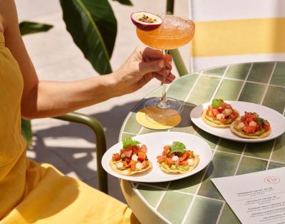 A person in a yellow outfit holds a tropical cocktail with a passion fruit garnish. Three small plates of appetizers sit on a green tiled table, creating a fresh, summery vibe.