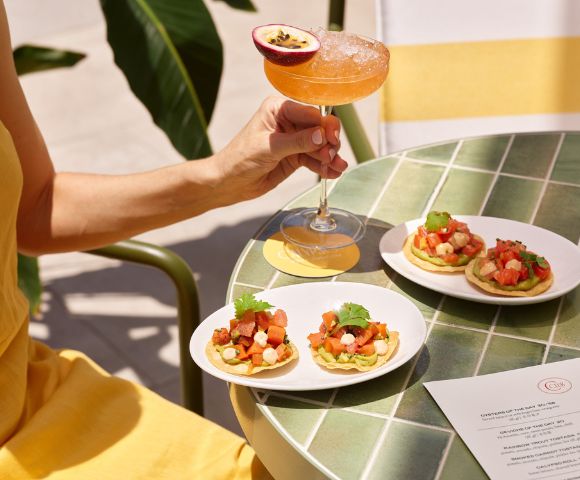A person in a yellow outfit holds a tropical cocktail with a passion fruit garnish. Three small plates of appetizers sit on a green tiled table, creating a fresh, summery vibe.