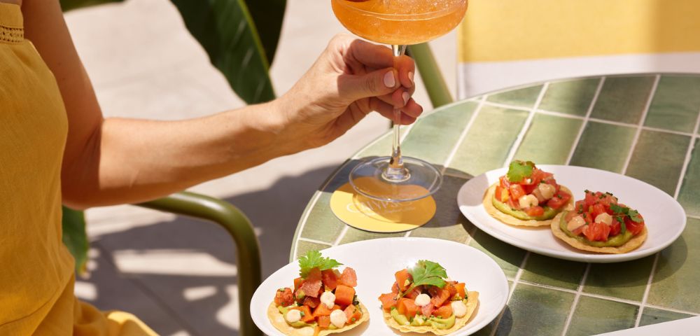 A person in a yellow outfit holds a tropical cocktail with a passion fruit garnish. Three small plates of appetizers sit on a green tiled table, creating a fresh, summery vibe.