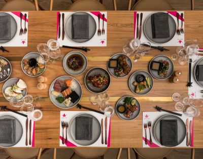 Elegant dining setup with a wooden table set for eight. Plates have gray napkins, accompanied by forks, knives, and chopsticks. Dishes of colorful food in the center create a vibrant contrast.