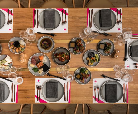 Elegant dining setup with a wooden table set for eight. Plates have gray napkins, accompanied by forks, knives, and chopsticks. Dishes of colorful food in the center create a vibrant contrast.