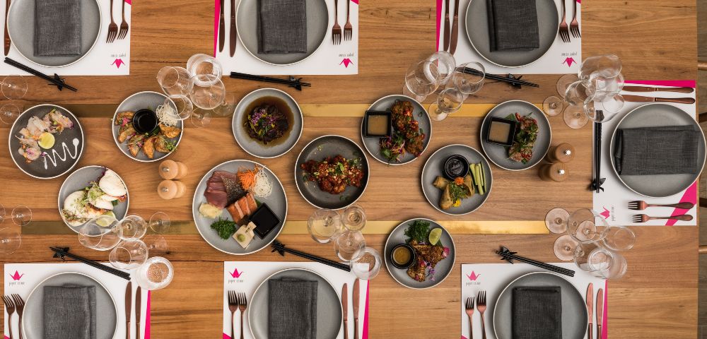 Elegant dining setup with a wooden table set for eight. Plates have gray napkins, accompanied by forks, knives, and chopsticks. Dishes of colorful food in the center create a vibrant contrast.
