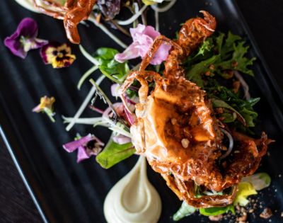 Gourmet dish featuring a golden, crispy soft-shell crab atop fresh greens, garnished with vibrant edible flowers on a sleek black plate.