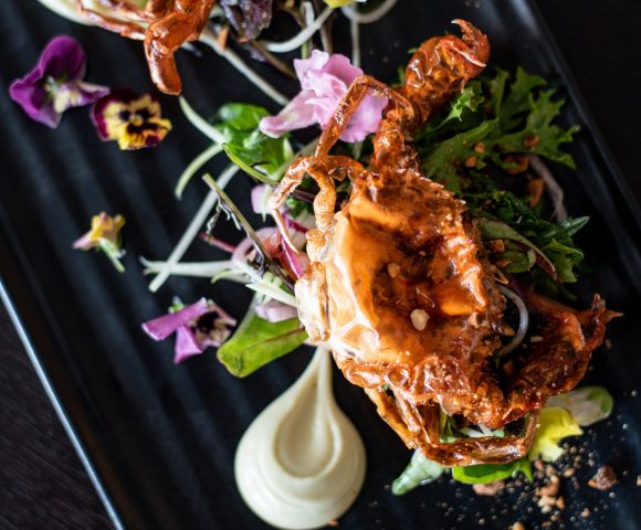 Gourmet dish featuring a golden, crispy soft-shell crab atop fresh greens, garnished with vibrant edible flowers on a sleek black plate.