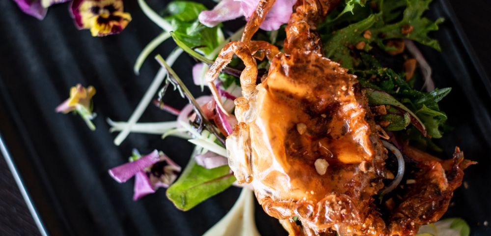 Gourmet dish featuring a golden, crispy soft-shell crab atop fresh greens, garnished with vibrant edible flowers on a sleek black plate.