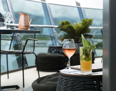 A stylish outdoor patio setting with a glass of rosé and a mojito on a wicker table. Background features modern railing, plants, and distant mountains.