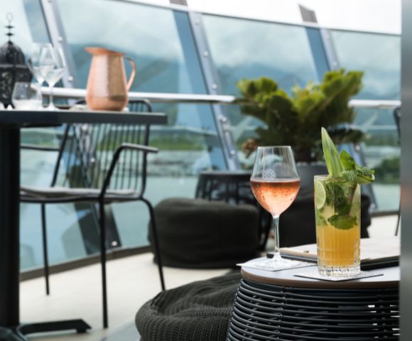 A stylish outdoor patio setting with a glass of rosé and a mojito on a wicker table. Background features modern railing, plants, and distant mountains.