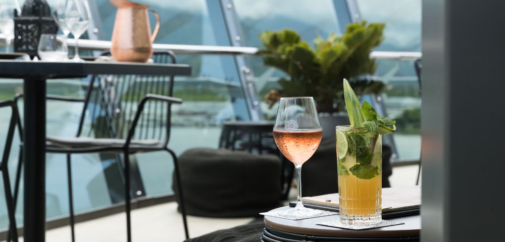 A stylish outdoor patio setting with a glass of rosé and a mojito on a wicker table. Background features modern railing, plants, and distant mountains.