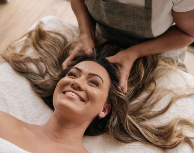 Woman smiling while receiving a relaxing head massage, lying on a white towel. Her long hair is spread out, and the atmosphere is calm and soothing.