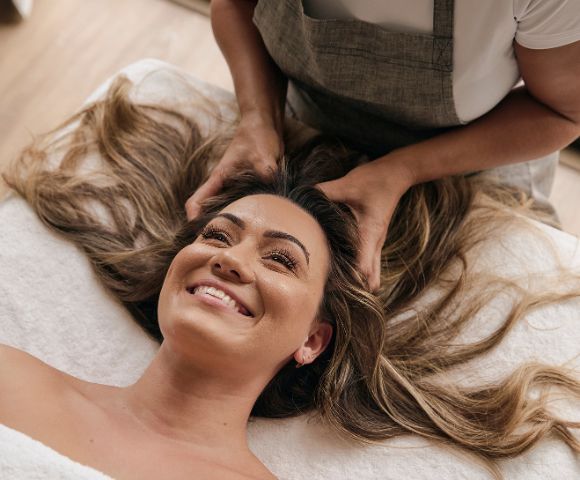 Woman smiling while receiving a relaxing head massage, lying on a white towel. Her long hair is spread out, and the atmosphere is calm and soothing.