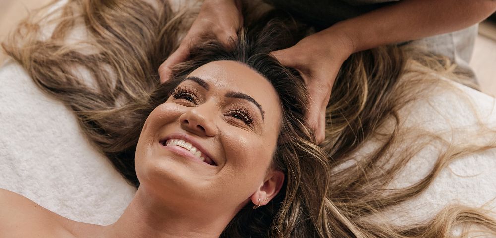 Woman smiling while receiving a relaxing head massage, lying on a white towel. Her long hair is spread out, and the atmosphere is calm and soothing.