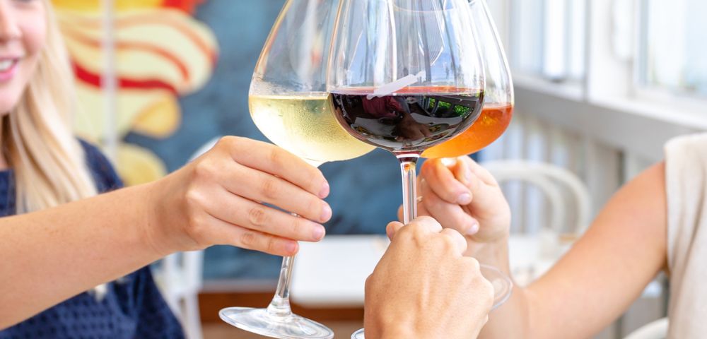 Three people clink glasses of red, white, and rosé wine in a bright setting, conveying a joyful, celebratory atmosphere.