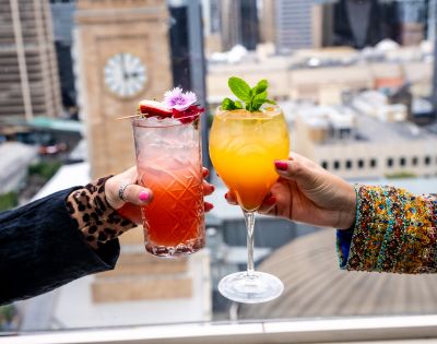 Two hands holding vibrant cocktails against a city skyline. One drink is red with floral garnish, the other orange with mint leaves; a clock tower is blurred in the background.