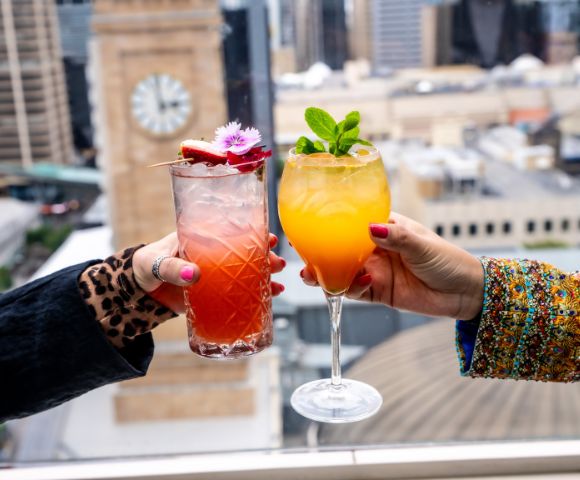 Two hands holding vibrant cocktails against a city skyline. One drink is red with floral garnish, the other orange with mint leaves; a clock tower is blurred in the background.
