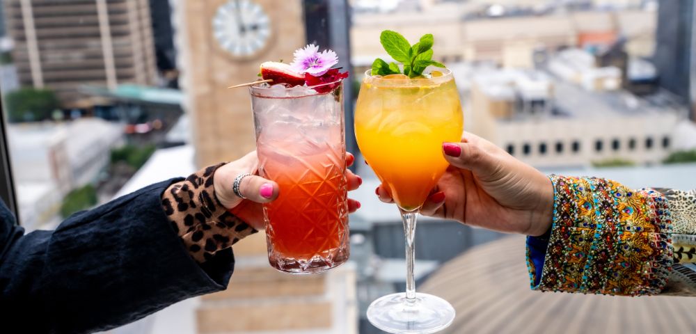 Two hands holding vibrant cocktails against a city skyline. One drink is red with floral garnish, the other orange with mint leaves; a clock tower is blurred in the background.