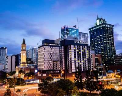 City skyline at dusk with illuminated modern skyscrapers and a historic clock tower. The sky is a gradient of blue, creating a vibrant urban scene.