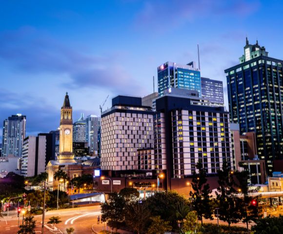 City skyline at dusk with illuminated modern skyscrapers and a historic clock tower. The sky is a gradient of blue, creating a vibrant urban scene.