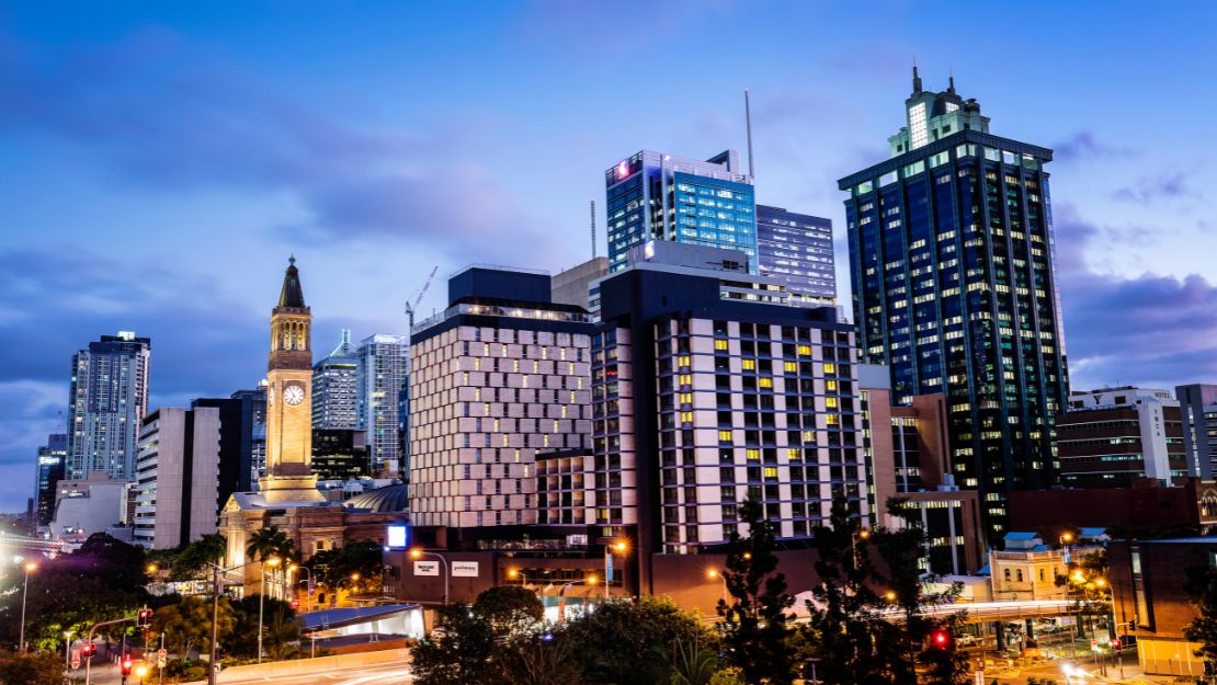 City skyline at dusk with illuminated modern skyscrapers and a historic clock tower. The sky is a gradient of blue, creating a vibrant urban scene.