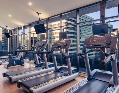 Modern gym with treadmills facing large windows. Cityscape visible outside, creating a sleek, motivated atmosphere. Treadmills' screens are illuminated.