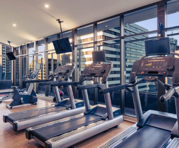 Modern gym with treadmills facing large windows. Cityscape visible outside, creating a sleek, motivated atmosphere. Treadmills' screens are illuminated.