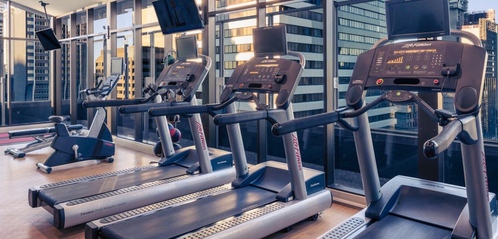 Modern gym with treadmills facing large windows. Cityscape visible outside, creating a sleek, motivated atmosphere. Treadmills' screens are illuminated.