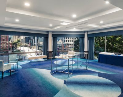 Modern conference room with large windows overlooking a cityscape. The space features blue patterned carpet, chairs, tables with refreshments, and a calm ambiance.