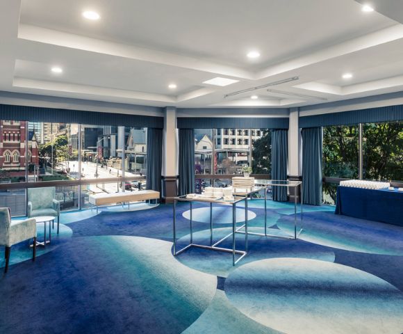 Modern conference room with large windows overlooking a cityscape. The space features blue patterned carpet, chairs, tables with refreshments, and a calm ambiance.