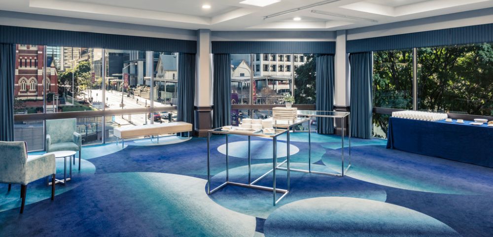 Modern conference room with large windows overlooking a cityscape. The space features blue patterned carpet, chairs, tables with refreshments, and a calm ambiance.