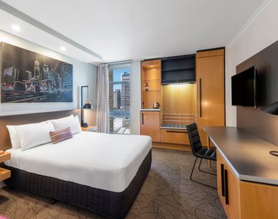 Modern hotel room with a neatly made bed, cityscape artwork, and large window offering a city view. Desk, chair, and wardrobe create a cozy ambiance.