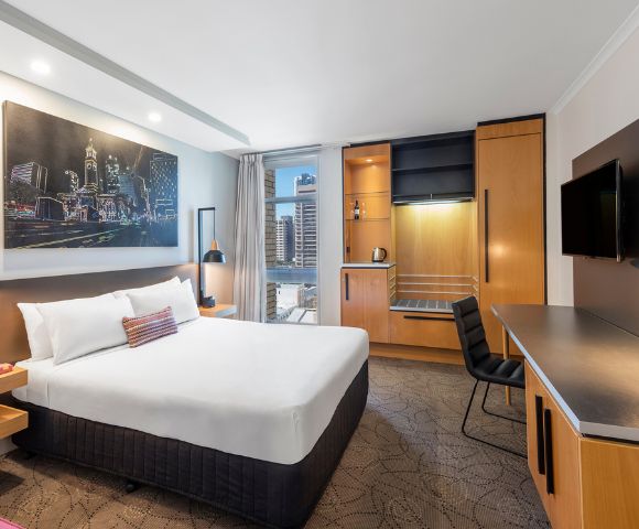 Modern hotel room with a neatly made bed, cityscape artwork, and large window offering a city view. Desk, chair, and wardrobe create a cozy ambiance.