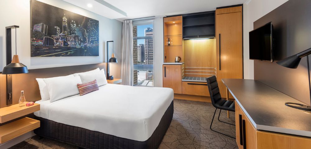Modern hotel room with a neatly made bed, cityscape artwork, and large window offering a city view. Desk, chair, and wardrobe create a cozy ambiance.