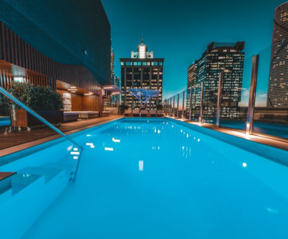 Sparkling rooftop pool at night, surrounded by modern skyscrapers. The water glows turquoise under a serene city skyline with ambient lighting.