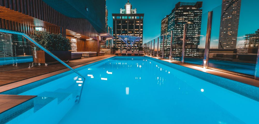 Sparkling rooftop pool at night, surrounded by modern skyscrapers. The water glows turquoise under a serene city skyline with ambient lighting.