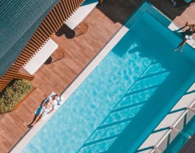 Aerial view of a serene poolside scene. A man relaxes on a sunbed beside a sparkling blue pool, surrounded by wooden decking. Shadows cast geometric patterns.