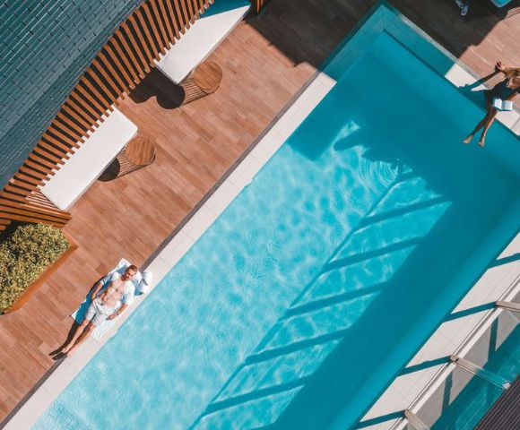 Aerial view of a serene poolside scene. A man relaxes on a sunbed beside a sparkling blue pool, surrounded by wooden decking. Shadows cast geometric patterns.