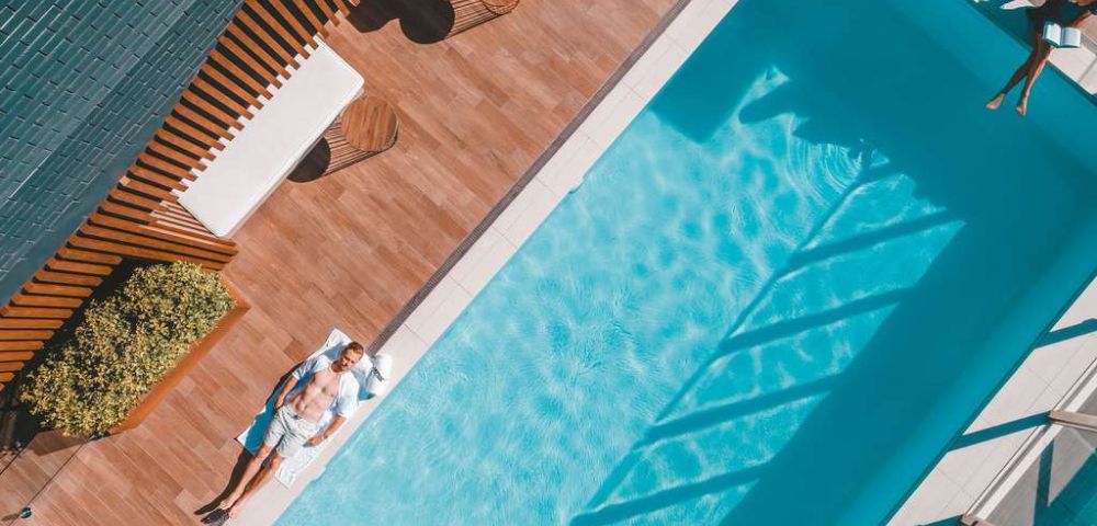 Aerial view of a serene poolside scene. A man relaxes on a sunbed beside a sparkling blue pool, surrounded by wooden decking. Shadows cast geometric patterns.