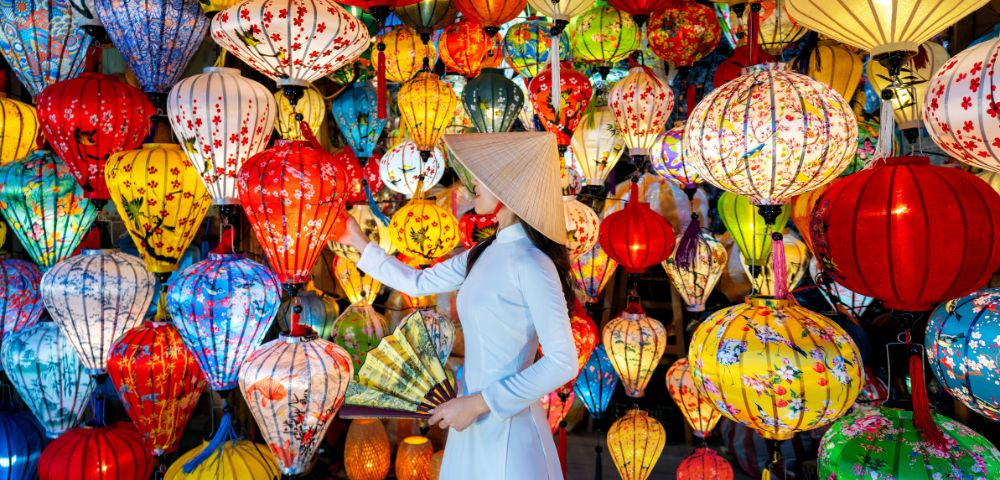 A woman wearing a conical hat admires a vibrant display of colorful, patterned lanterns in various shapes, evoking a festive and lively atmosphere.