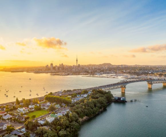 A scenic view of a city skyline at sunset, with a long bridge spanning a calm body of water. The sky is vibrant with golden hues, evoking tranquility.