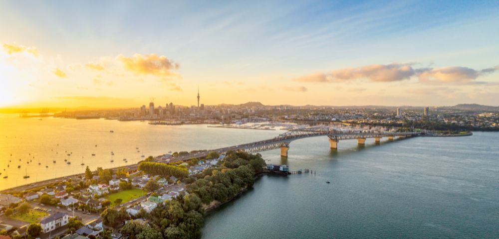 A scenic view of a city skyline at sunset, with a long bridge spanning a calm body of water. The sky is vibrant with golden hues, evoking tranquility.