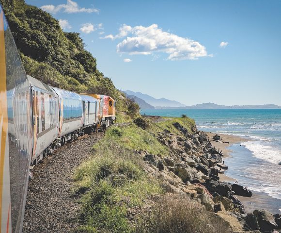A scenic train travels along a rocky coast, bordered by lush greenery and the sparkling blue sea. Mountains rise in the distance under a clear blue sky.