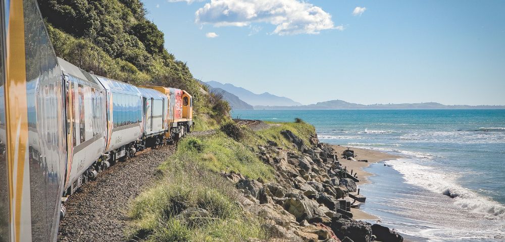 A scenic train travels along a rocky coast, bordered by lush greenery and the sparkling blue sea. Mountains rise in the distance under a clear blue sky.