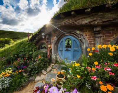 A picturesque hobbit house with a round blue door framed by vibrant flowers. Sunlight peeks through the clouds, casting a warm glow over the lush, grassy landscape.