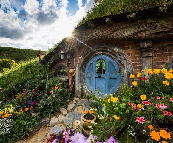 A picturesque hobbit house with a round blue door framed by vibrant flowers. Sunlight peeks through the clouds, casting a warm glow over the lush, grassy landscape.