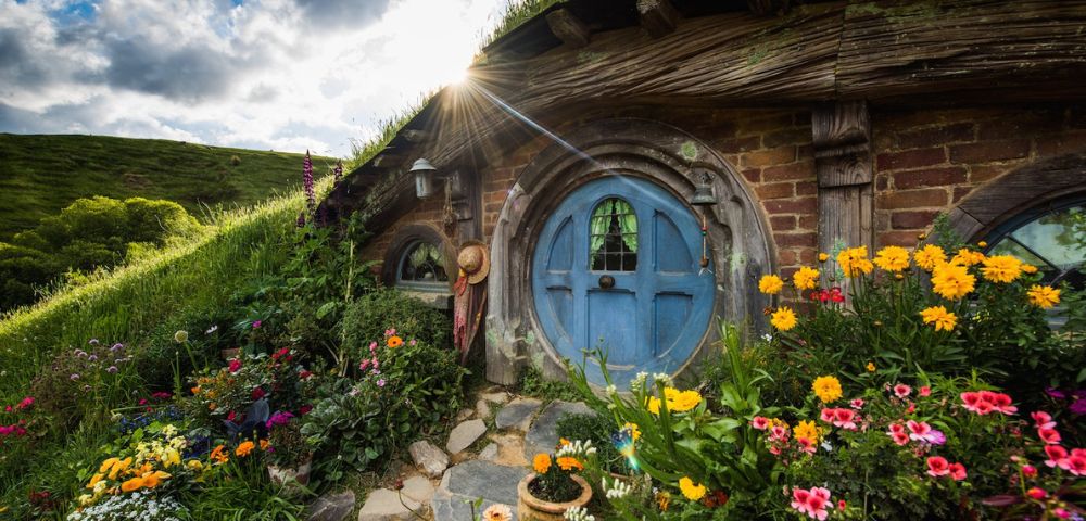 A picturesque hobbit house with a round blue door framed by vibrant flowers. Sunlight peeks through the clouds, casting a warm glow over the lush, grassy landscape.