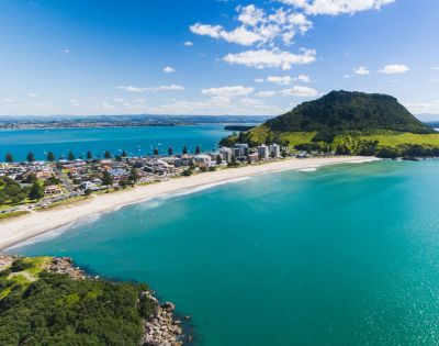 Aerial view of a coastal town with a prominent green mountain, a sandy beach, and turquoise sea. Blue sky with scattered clouds sets a serene tone.