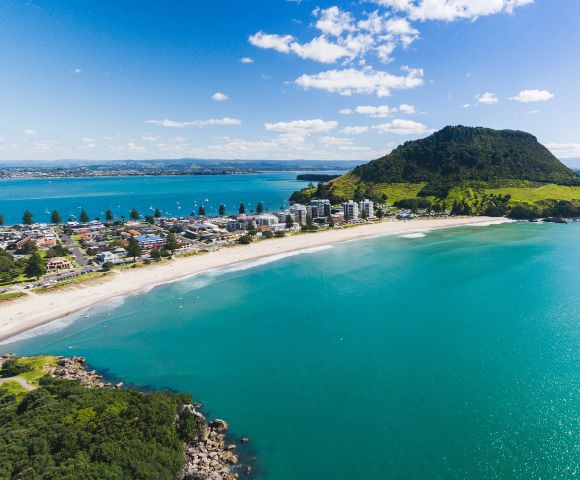 Aerial view of a coastal town with a prominent green mountain, a sandy beach, and turquoise sea. Blue sky with scattered clouds sets a serene tone.