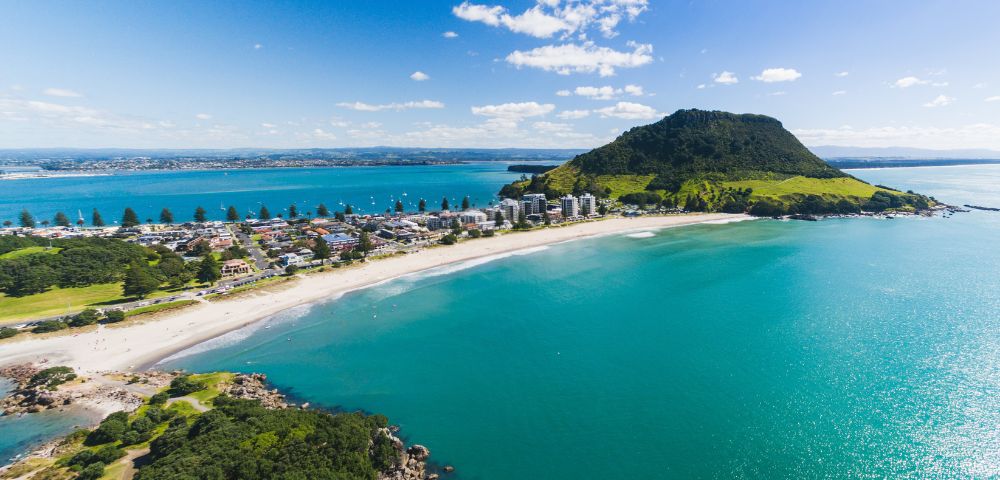 Aerial view of a coastal town with a prominent green mountain, a sandy beach, and turquoise sea. Blue sky with scattered clouds sets a serene tone.